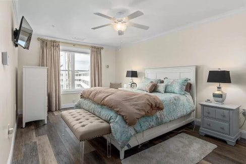 A bedroom with a double bed, floral bedding, a bench at the foot, a ceiling fan, a wall-mounted TV, and a window with curtains. Two nightstands with lamps are beside the bed.