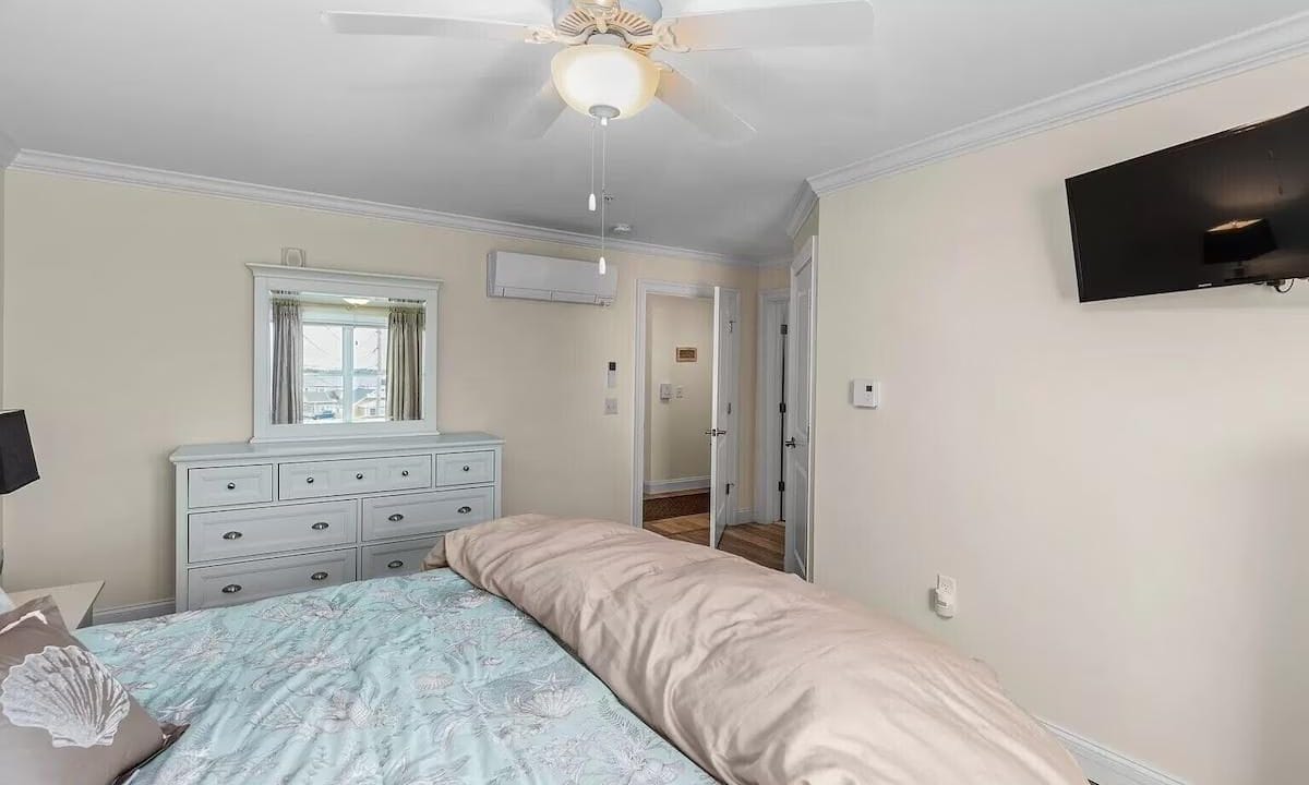 Bedroom with a ceiling fan, wall-mounted TV, dresser with a mirror, and a bed covered with a pastel comforter. Doorway visible leading to a hallway.