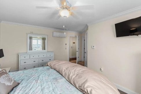 Bedroom with a ceiling fan, wall-mounted TV, dresser with a mirror, and a bed covered with a pastel comforter. Doorway visible leading to a hallway.