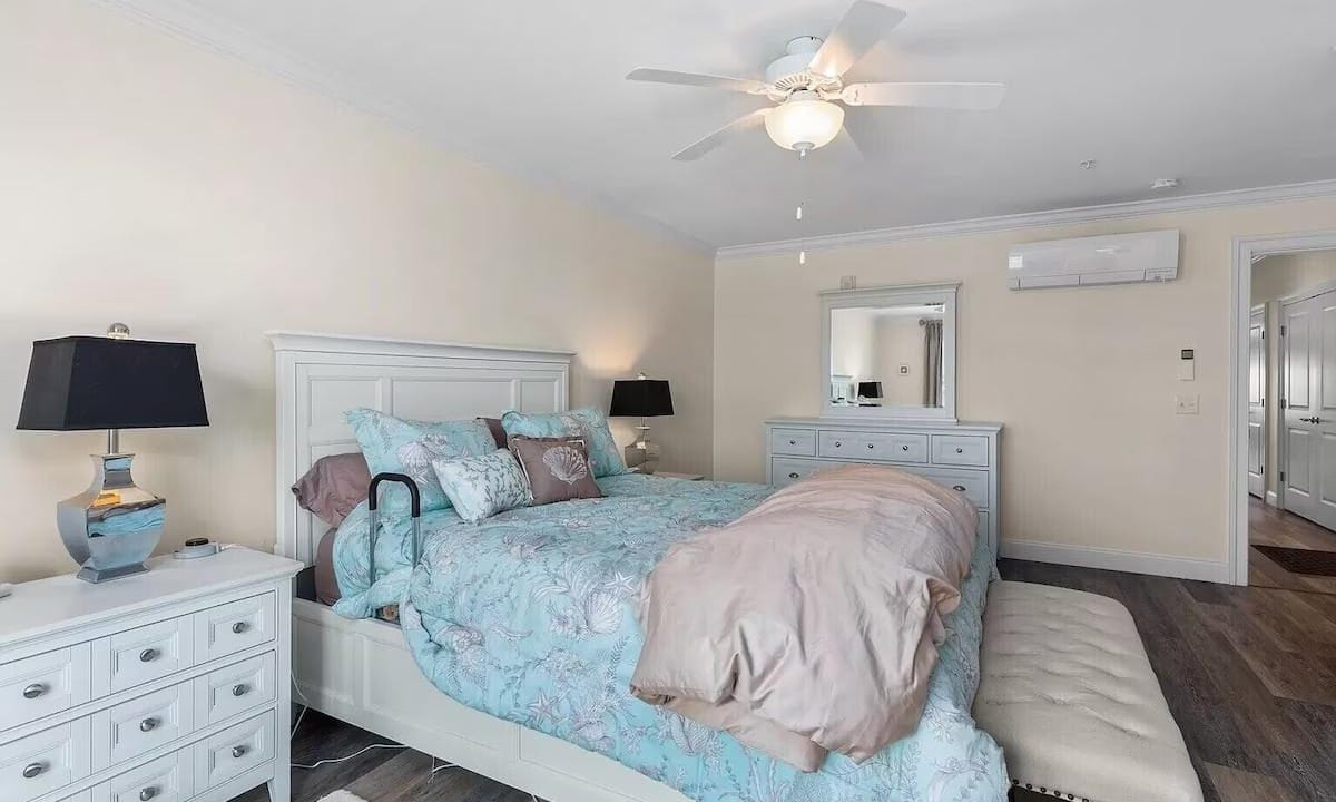 Bedroom with a bed featuring a floral duvet, nightstands with lamps, a dresser, a ceiling fan, and a mirror. Bench at the foot of the bed. Doorway visible in the background.