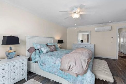 Bedroom with a bed featuring a floral duvet, nightstands with lamps, a dresser, a ceiling fan, and a mirror. Bench at the foot of the bed. Doorway visible in the background.