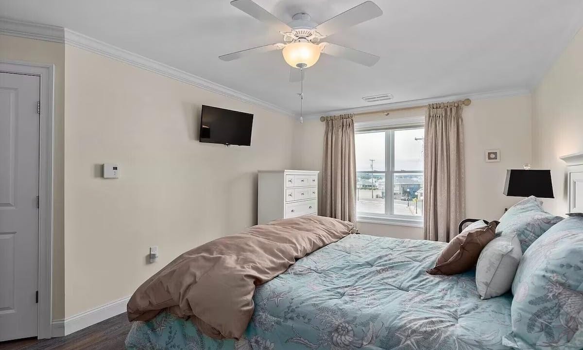 A bedroom with a floral bedspread, beige walls, TV mounted on the wall, and a window with beige curtains. A ceiling fan is above, and a dresser is near the window.