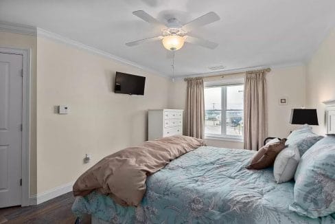 A bedroom with a floral bedspread, beige walls, TV mounted on the wall, and a window with beige curtains. A ceiling fan is above, and a dresser is near the window.