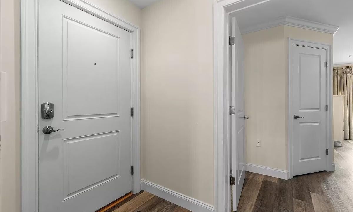 A hallway with three white doors: a main entrance on the left, an additional room on the right, and another visible in the background. The floor is wood laminate.