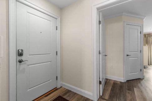 A hallway with three white doors: a main entrance on the left, an additional room on the right, and another visible in the background. The floor is wood laminate.