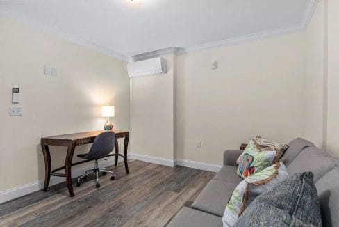 Room with a gray sofa, colorful cushions, wooden desk, chair, table lamp, and wall-mounted air conditioner. White walls and laminate flooring.