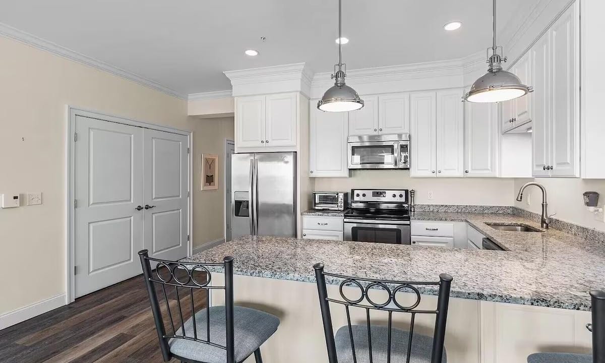 Modern kitchen with white cabinets, stainless steel appliances, granite countertops, and bar seating for two. Wooden flooring and pendant lights complete the look.