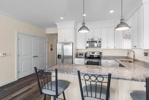 Modern kitchen with white cabinets, stainless steel appliances, granite countertops, and bar seating for two. Wooden flooring and pendant lights complete the look.