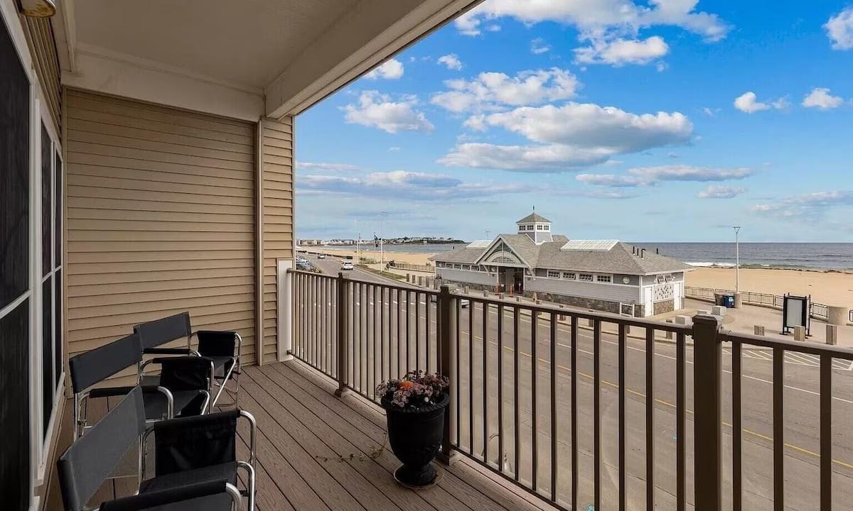 Balcony with black chairs overlooks a beachside road, building, and ocean horizon under a blue sky with clouds.