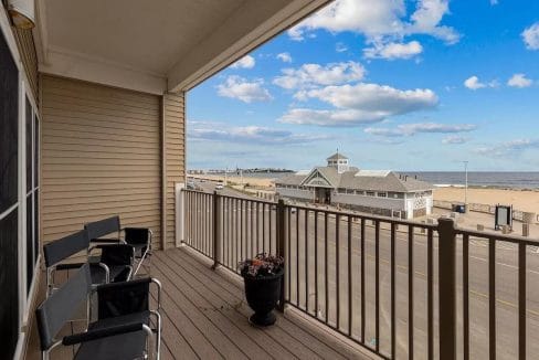 Balcony with black chairs overlooks a beachside road, building, and ocean horizon under a blue sky with clouds.