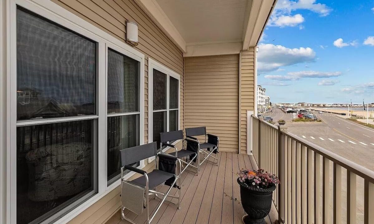 A balcony with three black chairs, two small tables, and a potted plant. Overlooks a street and ocean view on a clear day with scattered clouds.