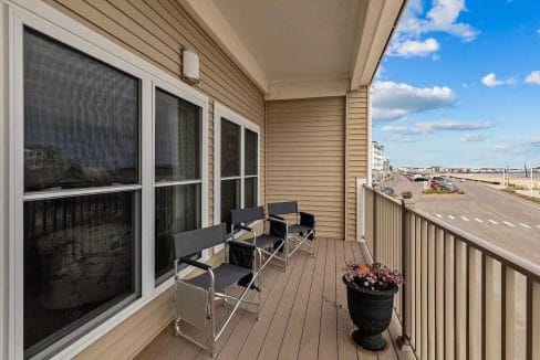 A balcony with three black chairs, two small tables, and a potted plant. Overlooks a street and ocean view on a clear day with scattered clouds.