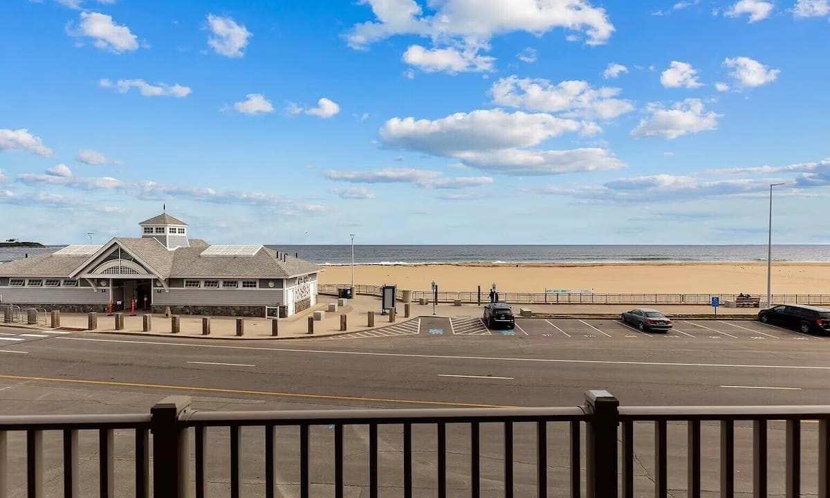 Beachfront scene with a boardwalk and parking area in the foreground, a pavilion to the left, and a clear blue sky with scattered clouds above.