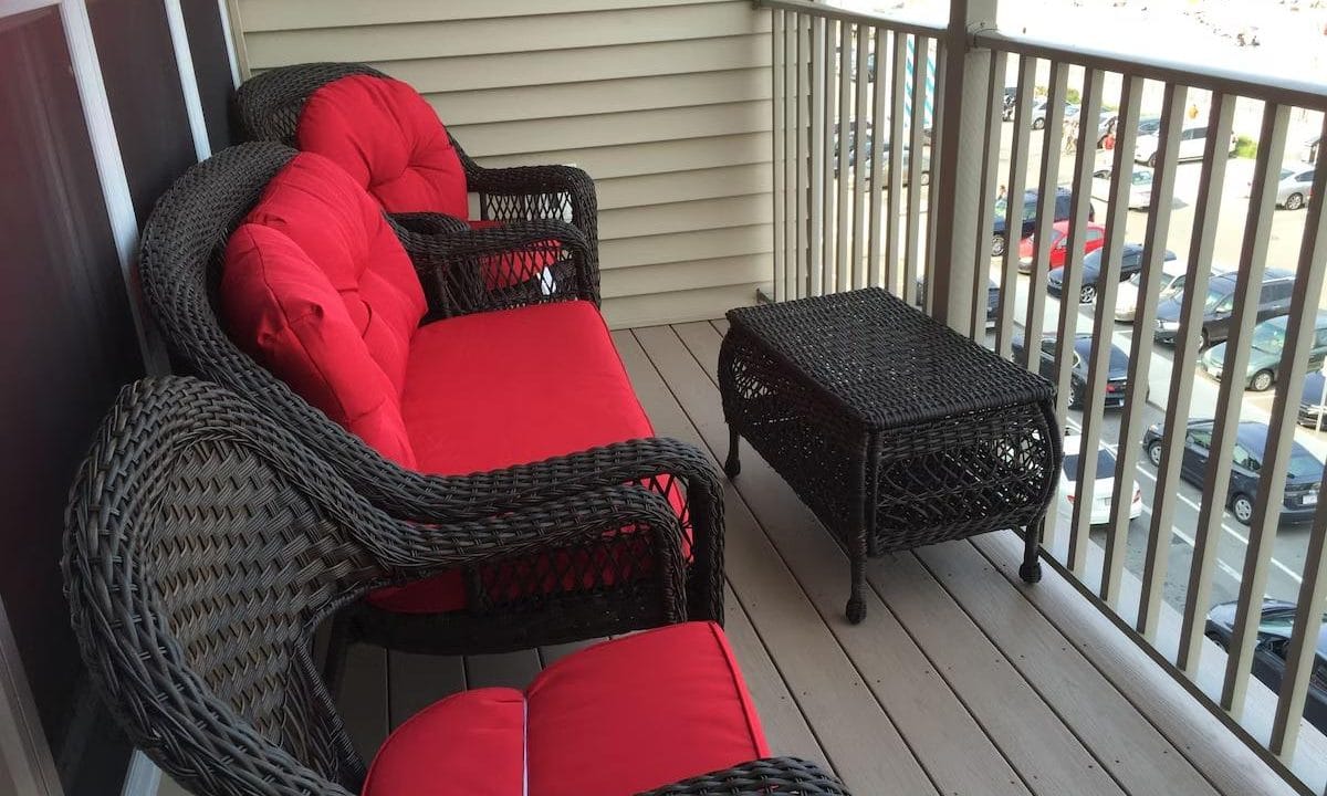 Wicker furniture with red cushions on a small balcony, overlooking a parking lot and partial view of the ocean.