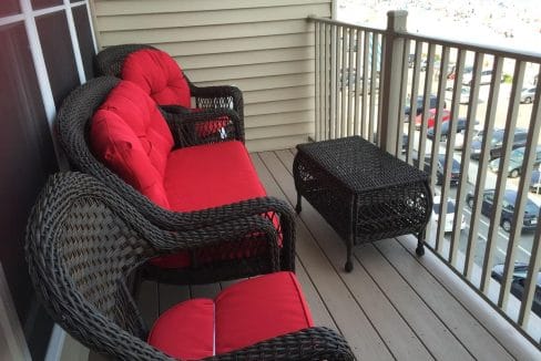 Wicker furniture with red cushions on a small balcony, overlooking a parking lot and partial view of the ocean.