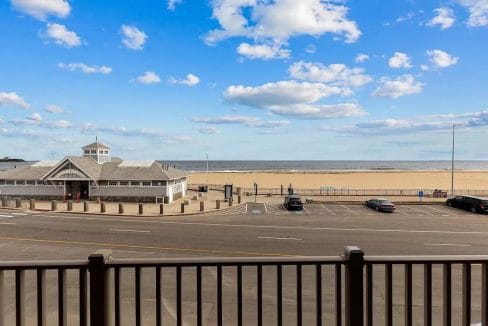 Beachfront scene with a boardwalk and parking area in the foreground, a pavilion to the left, and a clear blue sky with scattered clouds above.