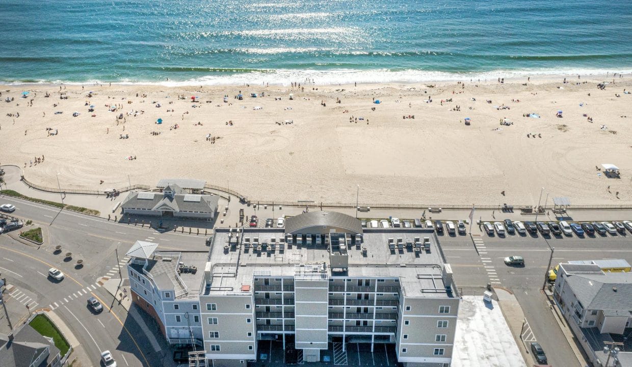 Aerial view of a beach with people, adjacent ocean, building, parking lot, and road intersection in the foreground.