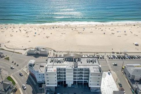 Aerial view of a beach with people, adjacent ocean, building, parking lot, and road intersection in the foreground.