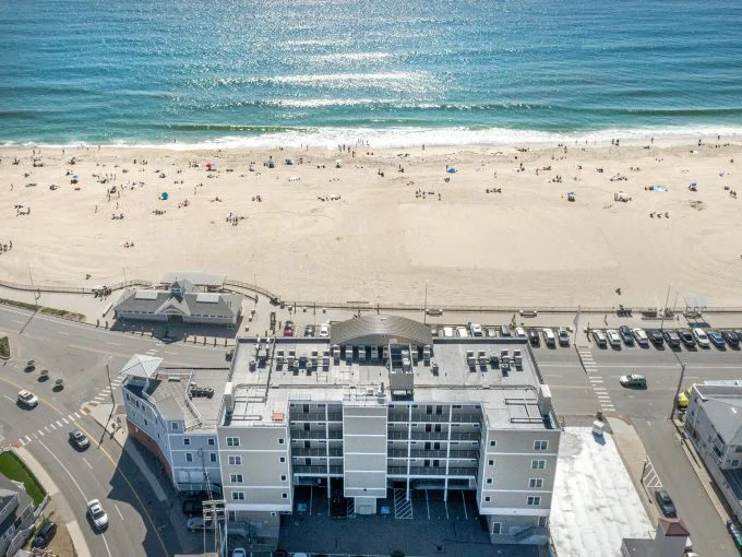 Aerial view of a beach with people, adjacent ocean, building, parking lot, and road intersection in the foreground.