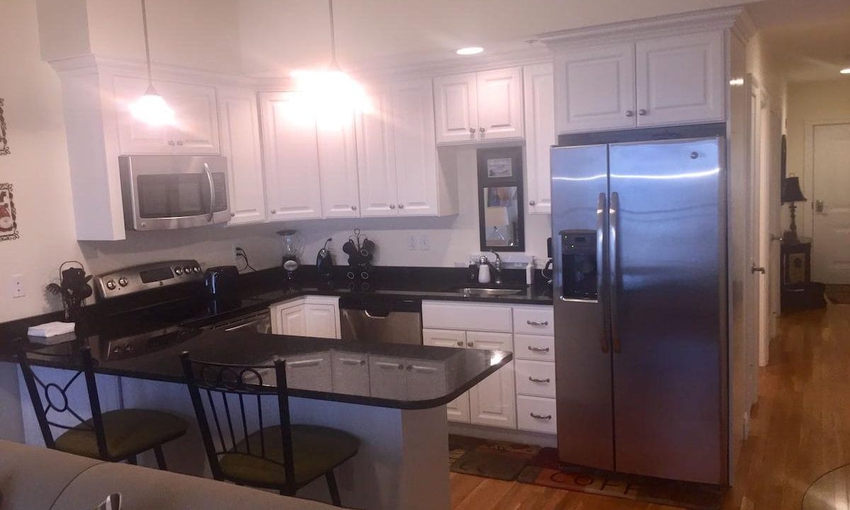 Modern kitchen with white cabinets, stainless steel appliances, black countertops, and a small island with two chairs. Wood flooring and ceiling lights are visible.