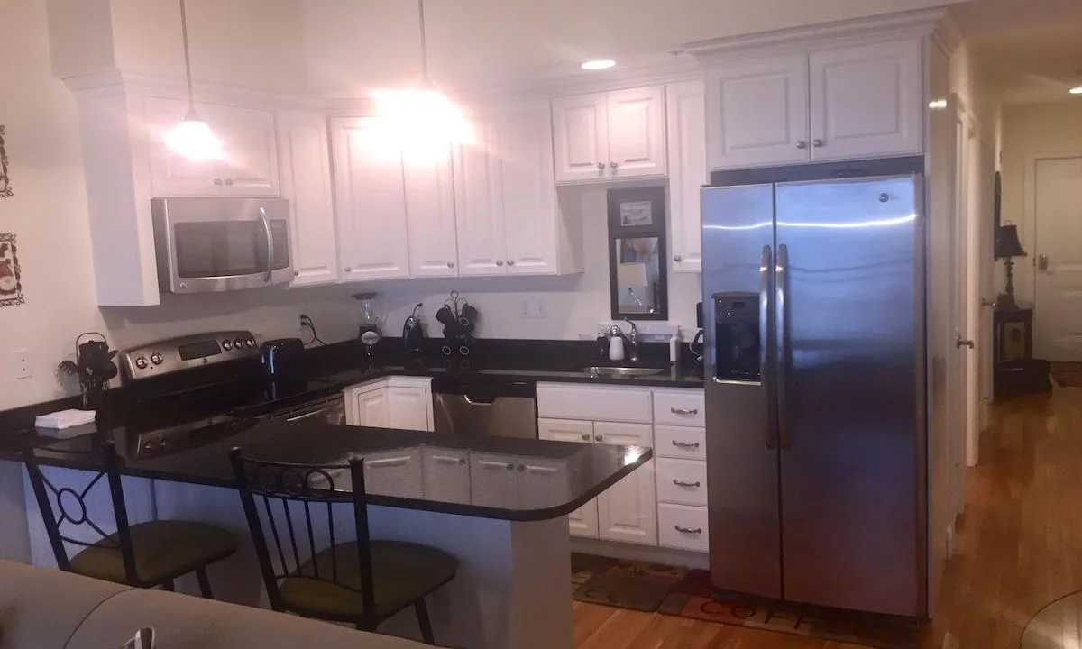 Modern kitchen with white cabinets, stainless steel appliances, black countertops, and a small island with two chairs. Wood flooring and ceiling lights are visible.