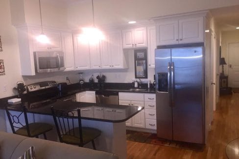 Modern kitchen with white cabinets, stainless steel appliances, black countertops, and a small island with two chairs. Wood flooring and ceiling lights are visible.