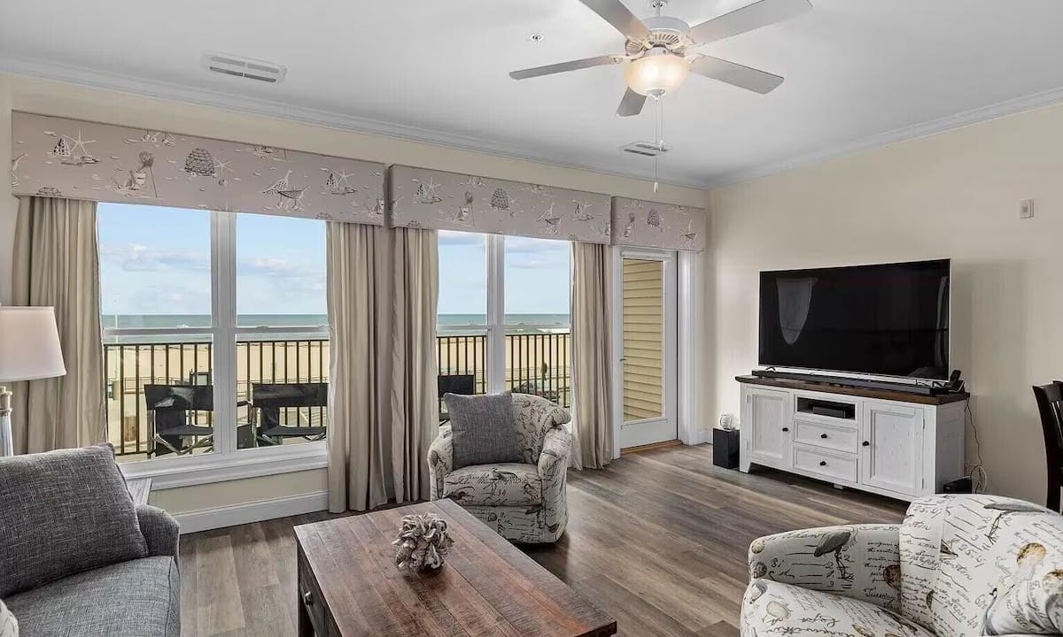 Living room with wood flooring, two armchairs, a sofa, coffee table, TV on a white stand, ceiling fan, and large windows showing a beach and ocean view.