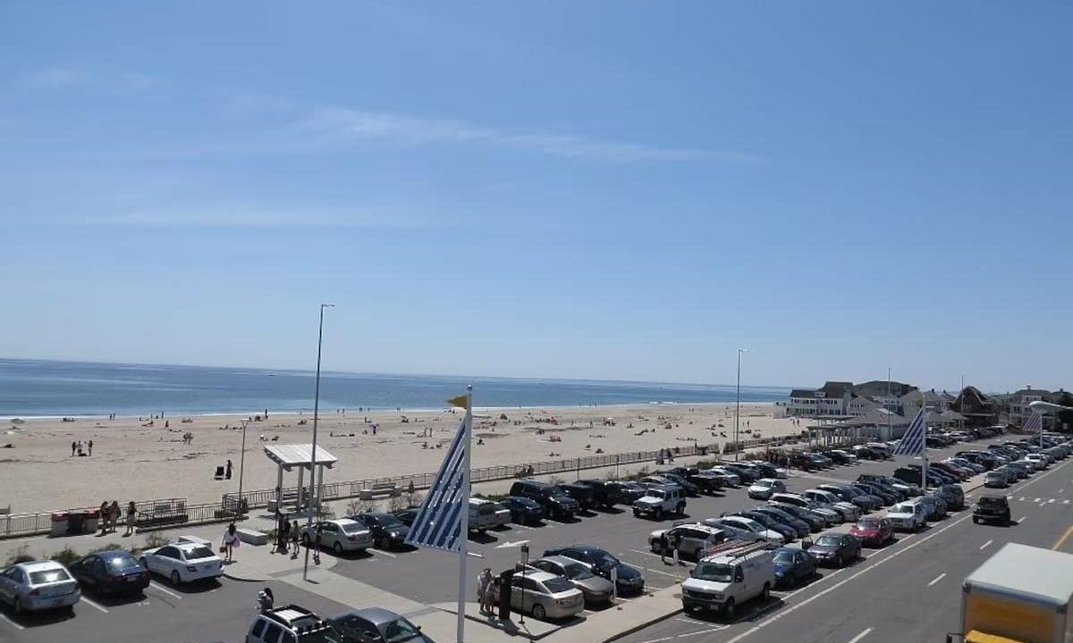 Beachfront scene with parked cars, sandy beach, and people by the ocean under a clear blue sky.