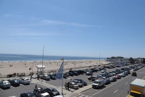 Beachfront scene with parked cars, sandy beach, and people by the ocean under a clear blue sky.