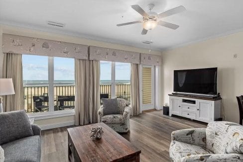 Living room with wood flooring, two armchairs, a sofa, coffee table, TV on a white stand, ceiling fan, and large windows showing a beach and ocean view.