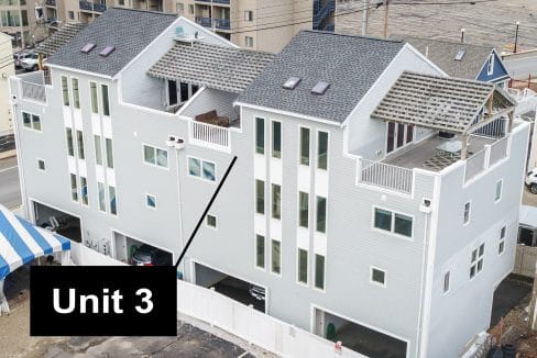 Aerial view of a row of three modern, pale gray townhouses with balconies. A black arrow points to the middle unit, labeled 