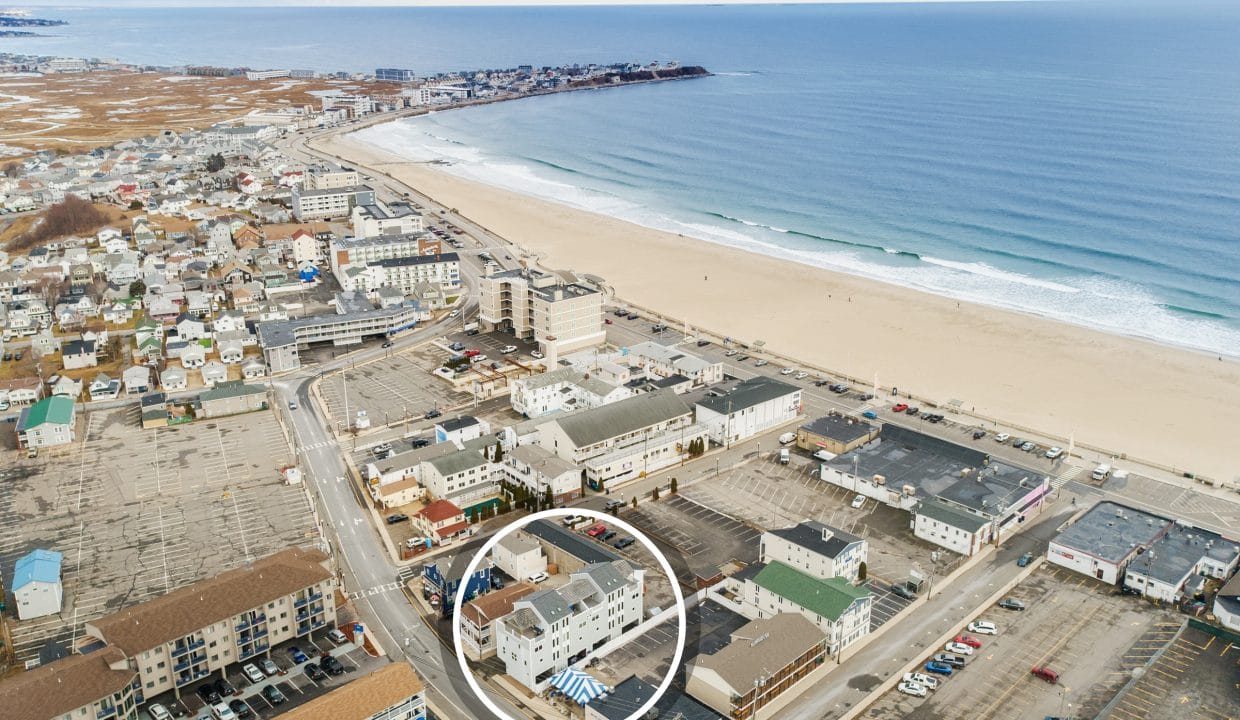 Aerial view of a coastal town with a long sandy beach. Buildings are arranged in a grid along streets. The ocean meets the shore with gentle waves. A single building is circled in white.