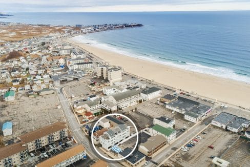 Aerial view of a coastal town with a long sandy beach. Buildings are arranged in a grid along streets. The ocean meets the shore with gentle waves. A single building is circled in white.