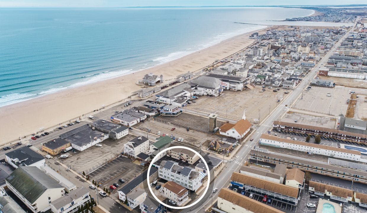 Aerial view of a coastal town with a beach and ocean on the left, and buildings and roads on the right. A building is circled in the foreground.