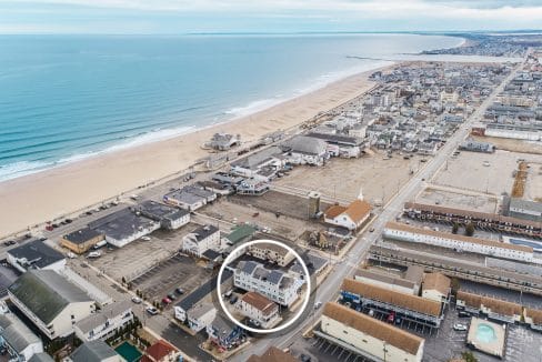 Aerial view of a coastal town with a beach and ocean on the left, and buildings and roads on the right. A building is circled in the foreground.