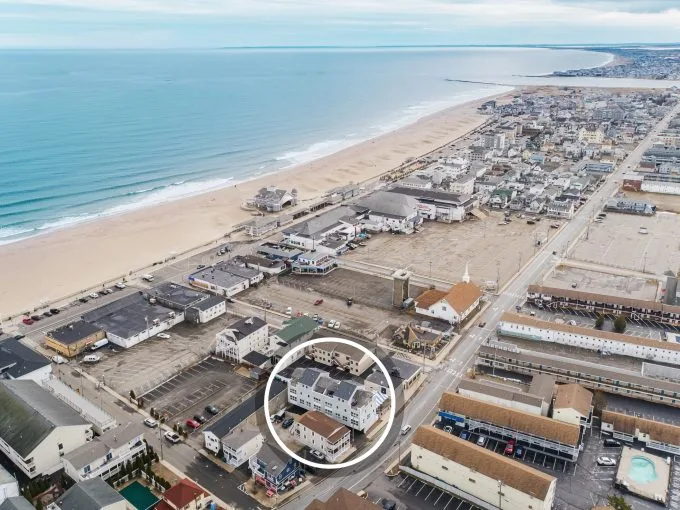 Aerial view of a coastal town with a beach and ocean on the left, and buildings and roads on the right. A building is circled in the foreground.