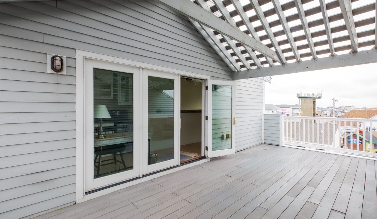 A rooftop deck with a wooden floor and pergola. Sliding glass doors lead inside, with a visible table and lamp. Nearby buildings and a lookout tower are seen in the background.