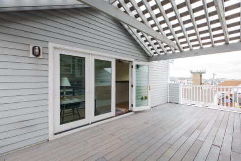 A rooftop deck with a wooden floor and pergola. Sliding glass doors lead inside, with a visible table and lamp. Nearby buildings and a lookout tower are seen in the background.