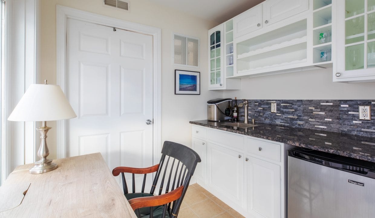 Small kitchenette with white cabinets, a dark backsplash, a wooden desk with a lamp, and a black chair. A mini fridge and coffee maker are present. A picture hangs on the wall.