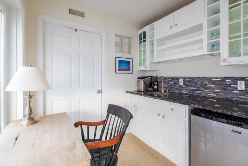 Small kitchenette with white cabinets, a dark backsplash, a wooden desk with a lamp, and a black chair. A mini fridge and coffee maker are present. A picture hangs on the wall.