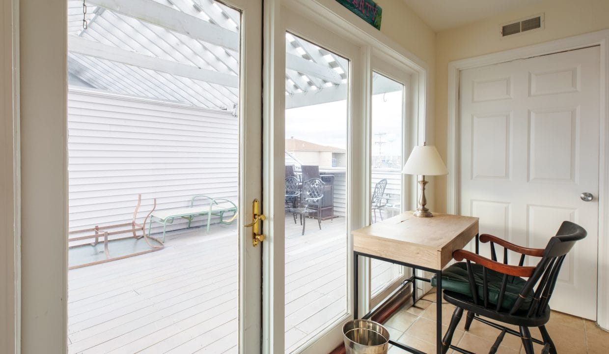 Room with glass doors leading to a patio. Inside, there's a wooden desk, chair, and a lamp. The patio has outdoor furniture and a pergola roof. A metal wastebasket is near the desk.