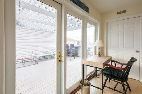 Room with glass doors leading to a patio. Inside, there's a wooden desk, chair, and a lamp. The patio has outdoor furniture and a pergola roof. A metal wastebasket is near the desk.