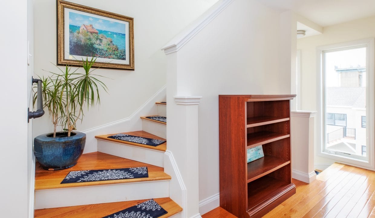 Wooden staircase with blue rugs leads to a landing with a bookshelf and potted plant. A framed painting hangs on the wall. Natural light fills the area from a nearby window.