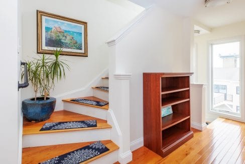 Wooden staircase with blue rugs leads to a landing with a bookshelf and potted plant. A framed painting hangs on the wall. Natural light fills the area from a nearby window.