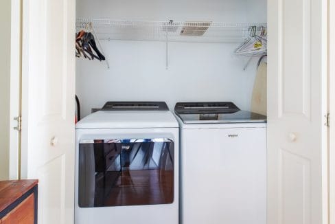 Laundry closet with white washing machine and dryer, shelf with hangers above, and an ironing board on the side.