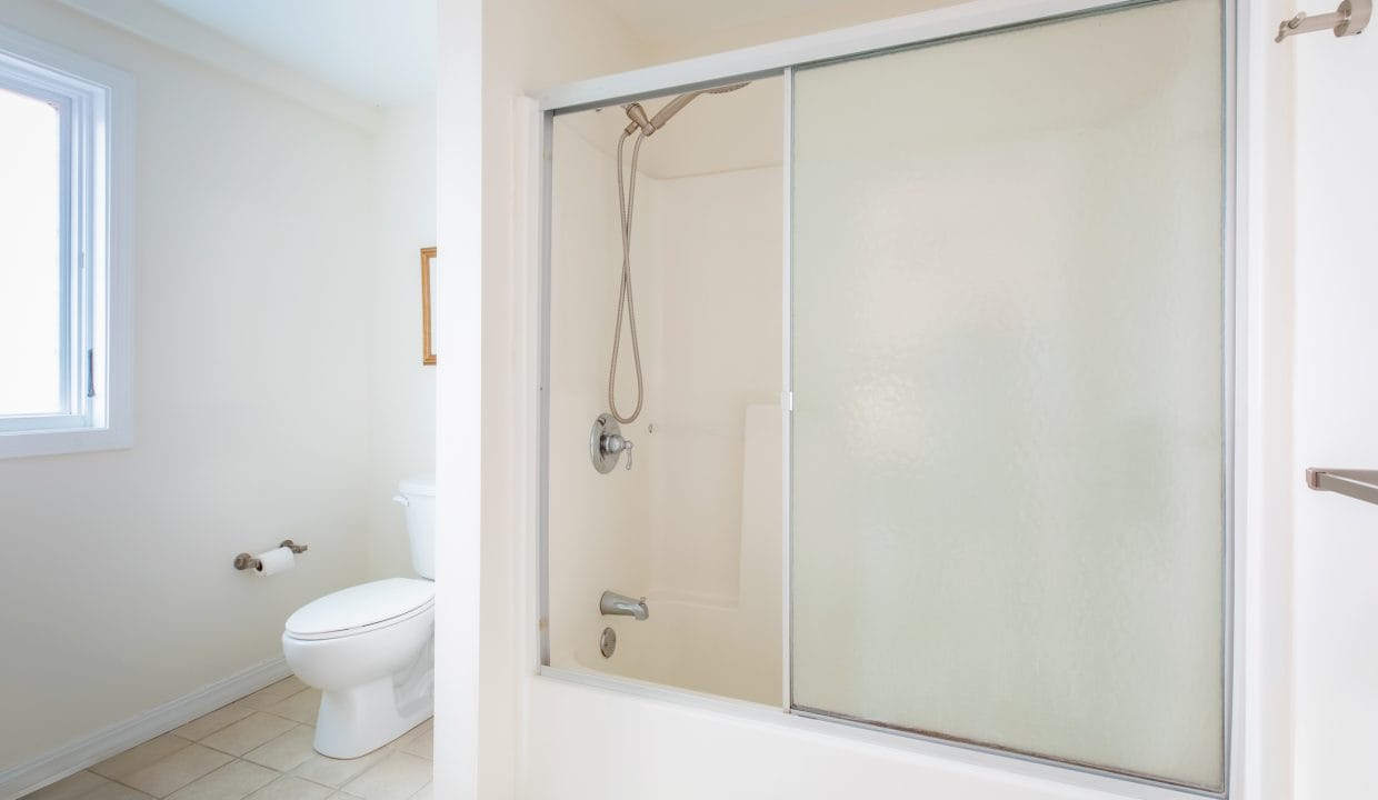 A bathroom with a shower enclosed by frosted sliding doors, a white toilet, and tiled floor. A window is on the left wall.