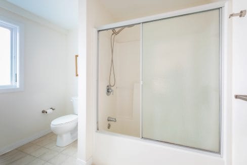 A bathroom with a shower enclosed by frosted sliding doors, a white toilet, and tiled floor. A window is on the left wall.