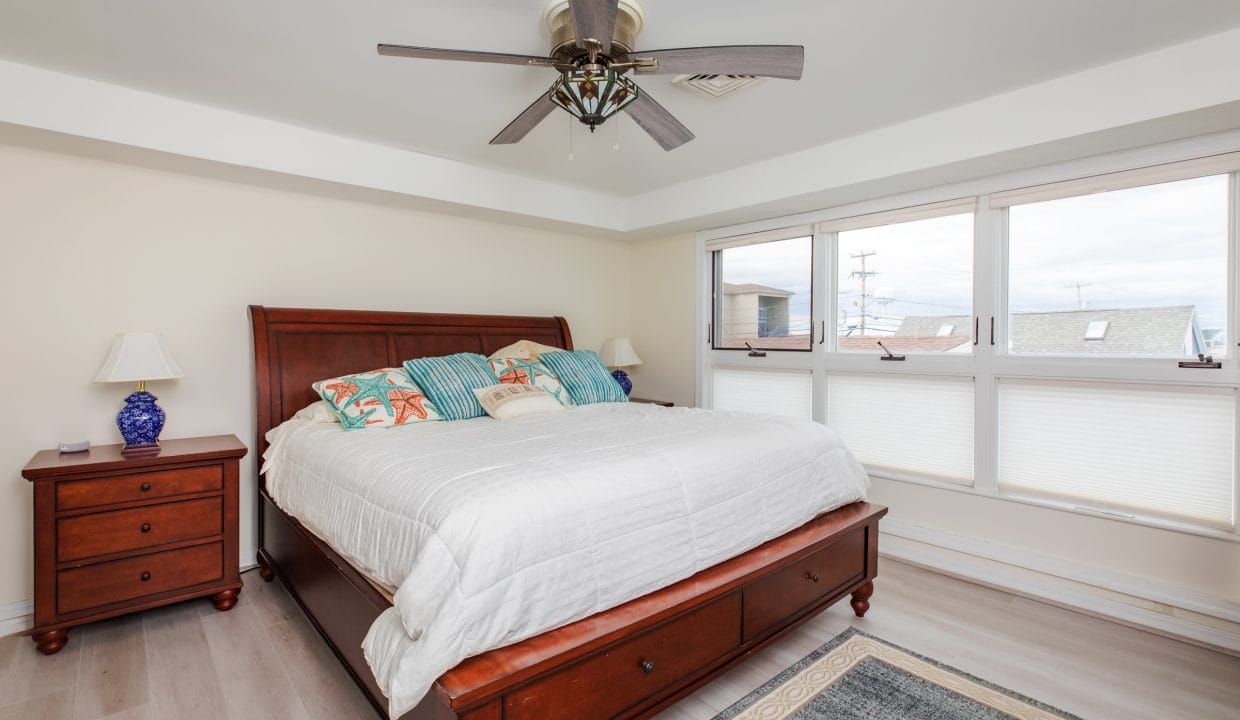 A bedroom with a large bed, colorful pillows, wooden nightstand with lamps, ceiling fan, and windows with a view.