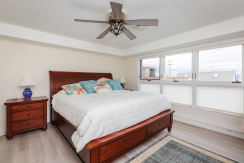 A bedroom with a large bed, colorful pillows, wooden nightstand with lamps, ceiling fan, and windows with a view.