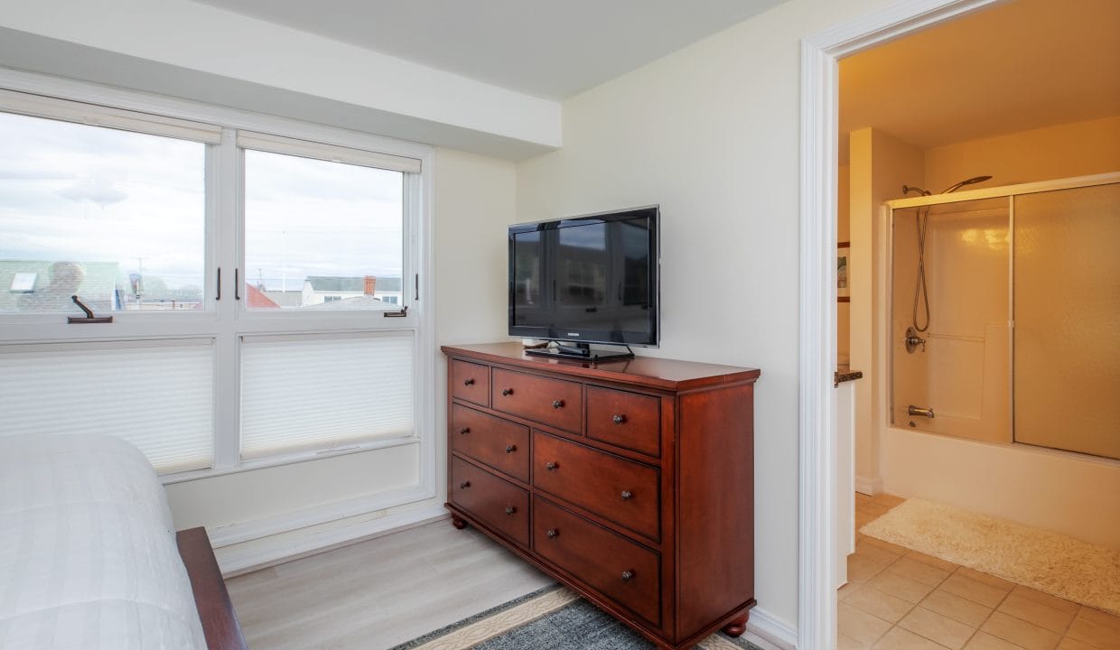 Bedroom with large window, chest of drawers with TV, and open door to a bathroom with a shower.